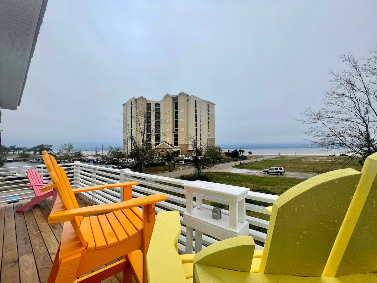 A large wrap-around patio features brightly colored Adirondack chairs in pink, yellow, and orange. The panoramic view includes a multi-story building in the distance with gray skies above and a glimpse of the beach and sea.