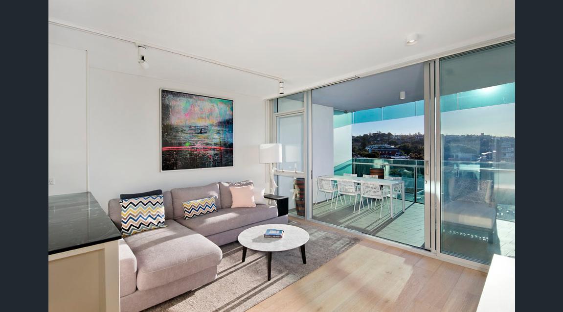 A modern living area features a spacious light-grey sofa adorned with patterned cushions, complemented by a round coffee table. A large abstract artwork decorates the wall. Floor-to-ceiling glass doors open to a balcony, allowing natural light and views into the room.