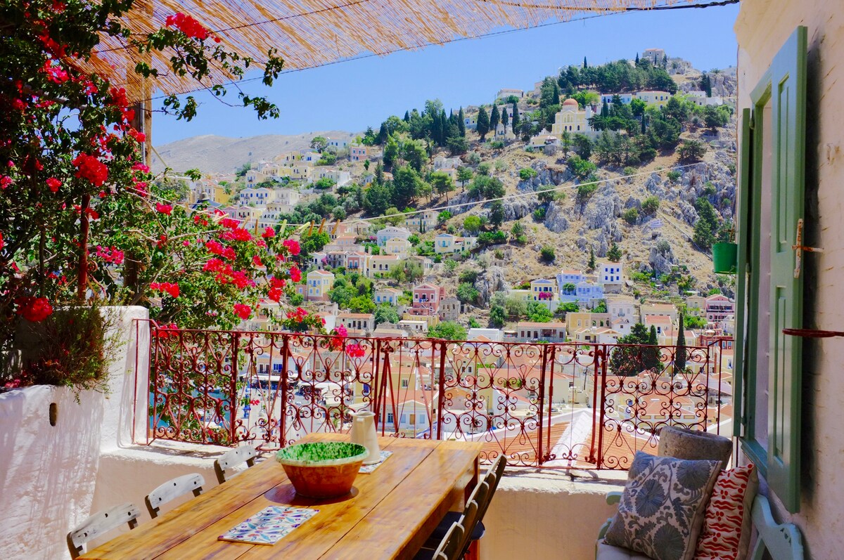 A large wooden table is adorned with a bowl and small decorative items, set under a shaded pergola. Vibrant flowers in shades of red bloom alongside the terrace railing, while a picturesque view of the hillside houses is captured in the background, showcasing the surrounding landscape.