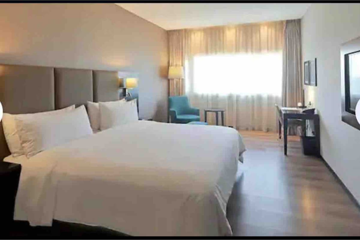 A spacious bedroom features a large bed with white linens, complemented by two bedside lamps. A window with sheer curtains allows natural light to fill the room. A chair and small table are positioned near the window, and a television is mounted on the wall.