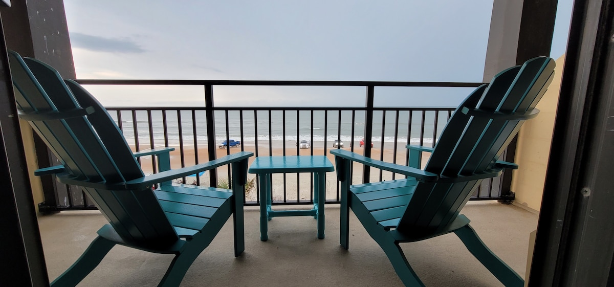 Two teal adirondack chairs are positioned on a balcony, facing the ocean. A small table sits between the chairs. The tranquil view includes rolling waves and a stretch of sandy beach, providing an inviting outdoor space for relaxation.