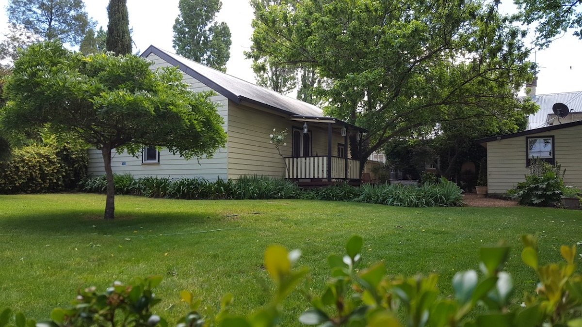 A charming single-story structure is framed by lush greenery and trees, with a welcoming porch and front steps leading to the entrance. The surrounding garden features manicured lawns and varied shrubs, enhancing the peaceful outdoor environment.
