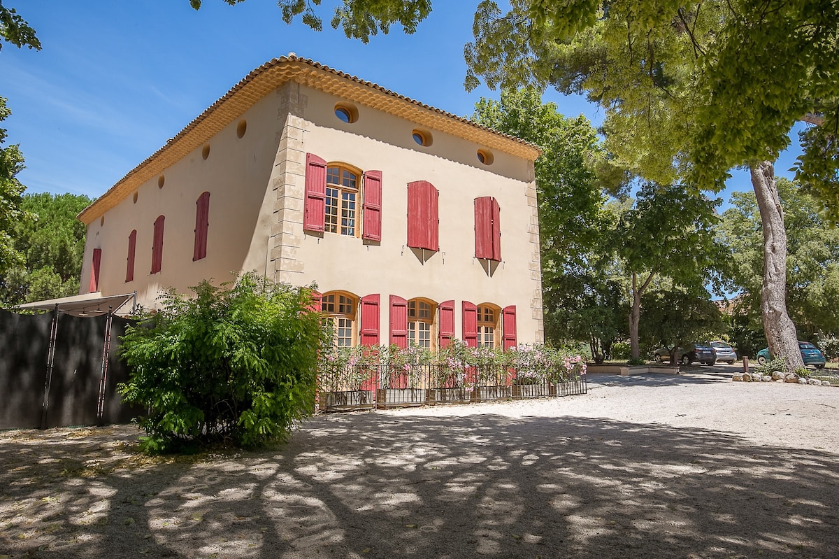 The exterior of a historic building is showcased, featuring warm-toned walls complemented by red shutters. Surrounding vegetation casts intriguing shadows on the gravel path, and vibrant greenery is visible against the structure. Ample windows illuminate the ground floor, enhancing the welcoming appearance.