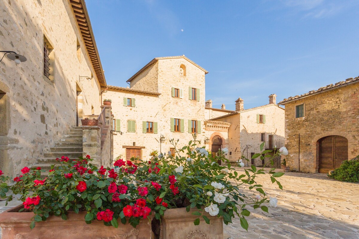 An outdoor view of a historical stone building features multiple windows with green shutters. Bright red and white flowers in pots adorn the foreground, while the surrounding landscape includes other rustic stone structures. The sky is clear with soft daylight illuminating the scene.
