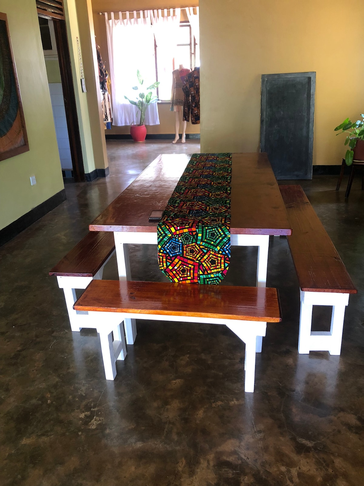 A large wooden dining table is accompanied by two matching benches, all set against a warm yellow wall. A colorful kente cloth is placed lengthwise on the table, providing a vibrant contrast to the natural wood. Natural light fills the space from nearby windows.