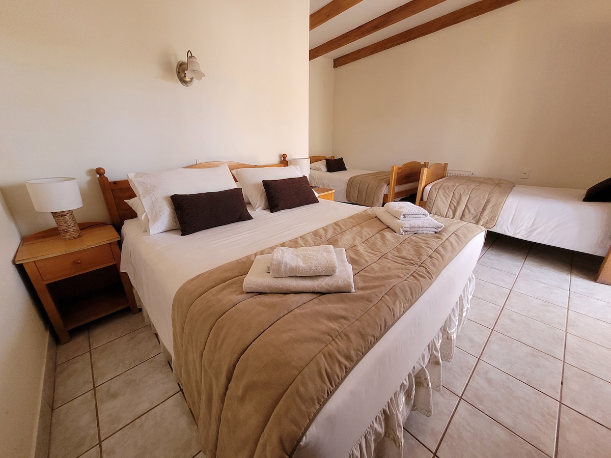 A cozy bedroom is equipped with a spacious double bed accompanied by two single beds. Soft linens are arranged neatly across the beds, and towels are placed atop the covers. Natural light fills the room, highlighting the wooden furniture and tiled flooring.