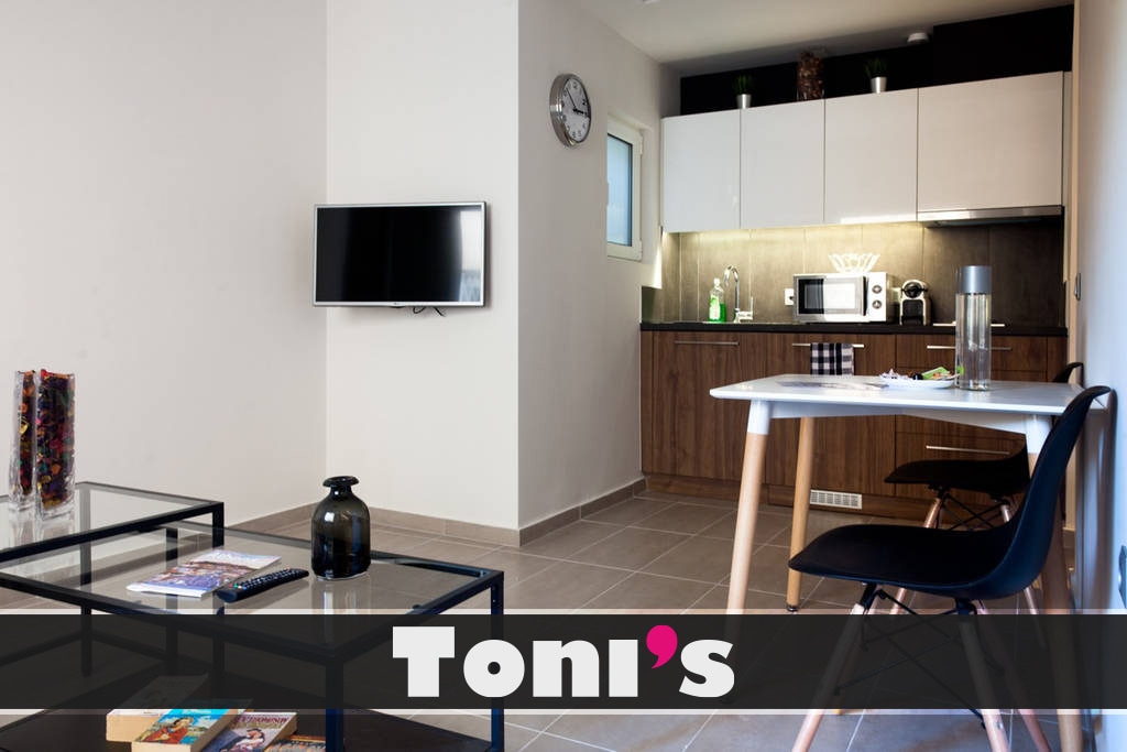 A modern kitchen area is visible, featuring sleek cabinetry and essential appliances. A small dining table with chairs is situated near a wall-mounted television. A glass coffee table in the foreground adds to the open space, complemented by a simple black vase.