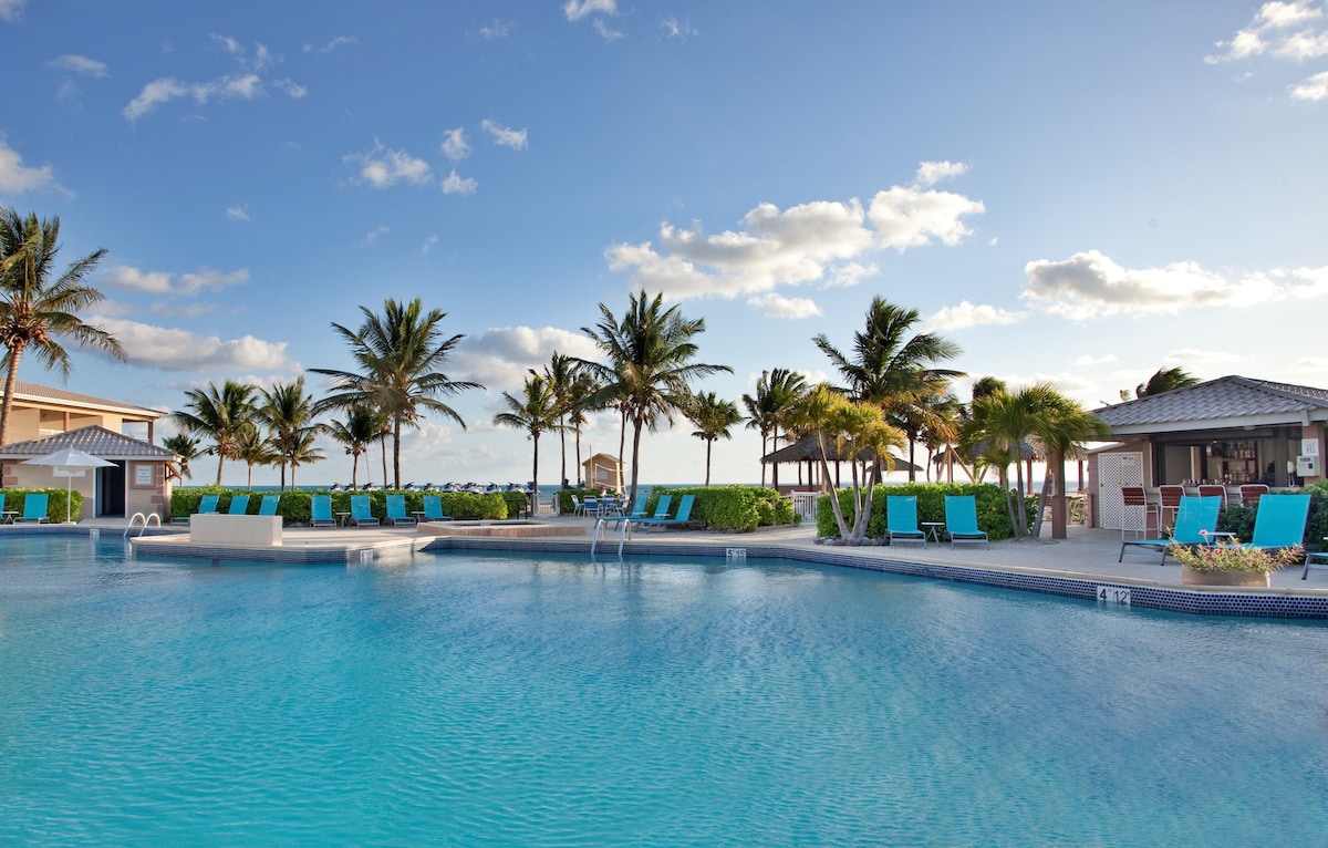 A large outdoor pool area is framed by lush palm trees and well-maintained greenery. Multiple lounge chairs are arranged along the water's edge, with a shaded gazebo visible in the background. The clear sky enhances the inviting atmosphere of the pool area.