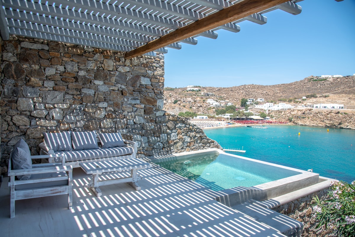 A private balcony offers a serene view of the sea, featuring a small swimming pool beside a comfortable seating area with striped cushions. Sunlight creates a pattern of shadows through the wooden beams overhead, illuminating the stone wall façade and the vibrant blue water below.