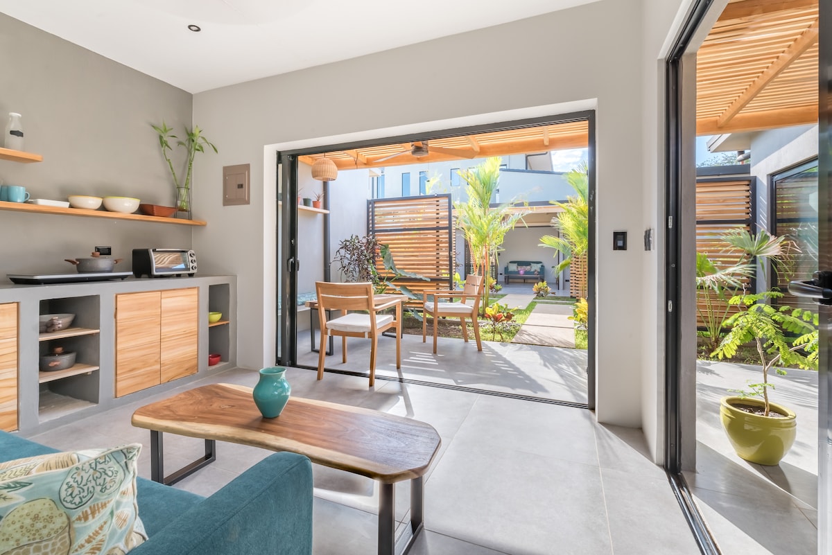 An open living area is visible featuring a seating arrangement with a blue couch and a wooden coffee table. Large sliding glass doors connect the space to an outdoor patio with chairs and potted plants, allowing natural light to fill the room.