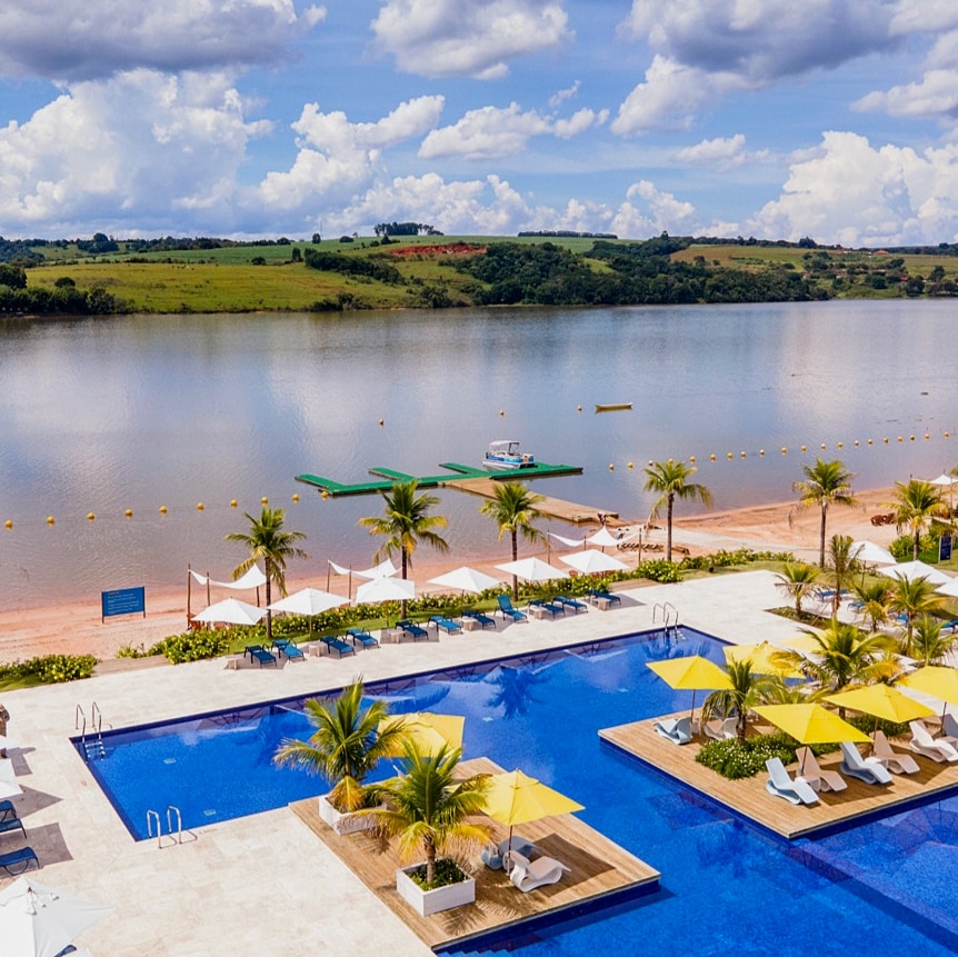 A tranquil view of a large swimming pool adjacent to a sandy beach and calm water. Sun loungers with vibrant yellow umbrellas line the poolside, while palm trees provide gentle shade. In the distance, a clear view of the water and green landscape is visible.