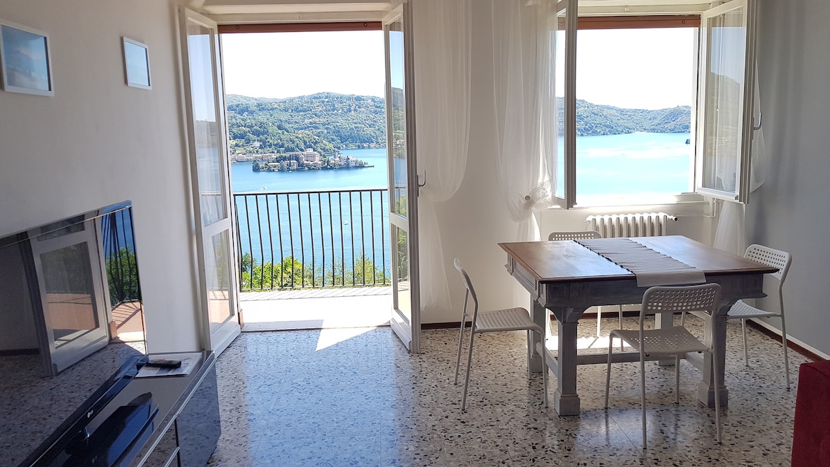 A bright living area features large open windows that frame a panoramic view of Lake Orta and San Giulio Island. A wooden table with white chairs is positioned in front of the windows, and a television unit is placed against the opposite wall, enhancing the spacious atmosphere.