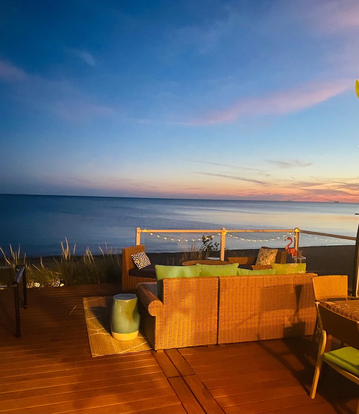 A comfortable seating area is showcased on a spacious deck, facing serene waters. Soft lighting illuminates the surroundings, while the sky exhibits gentle hues of blue and pink from the sunset. Beach grass frames the view, enhancing the coastal ambiance.