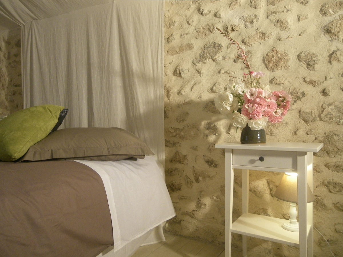 A cozy sleeping area is characterized by a bed with a combination of neutral and green pillows. A small white bedside table holds a vase with pink flowers, and a soft light is emitted from a lamp on the table, enhancing the stony wall backdrop.