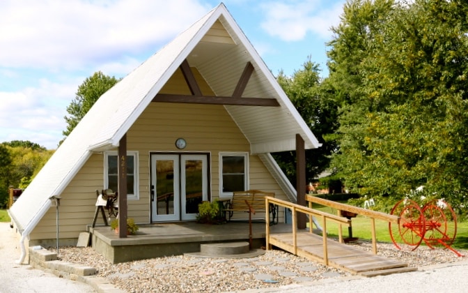 The A-frame exterior is showcased with a sloped roof and a welcoming front entrance. Large glass doors allow natural light into the space, while a small porch area is lined with seating. Surrounding greenery complements the tranquil setting.