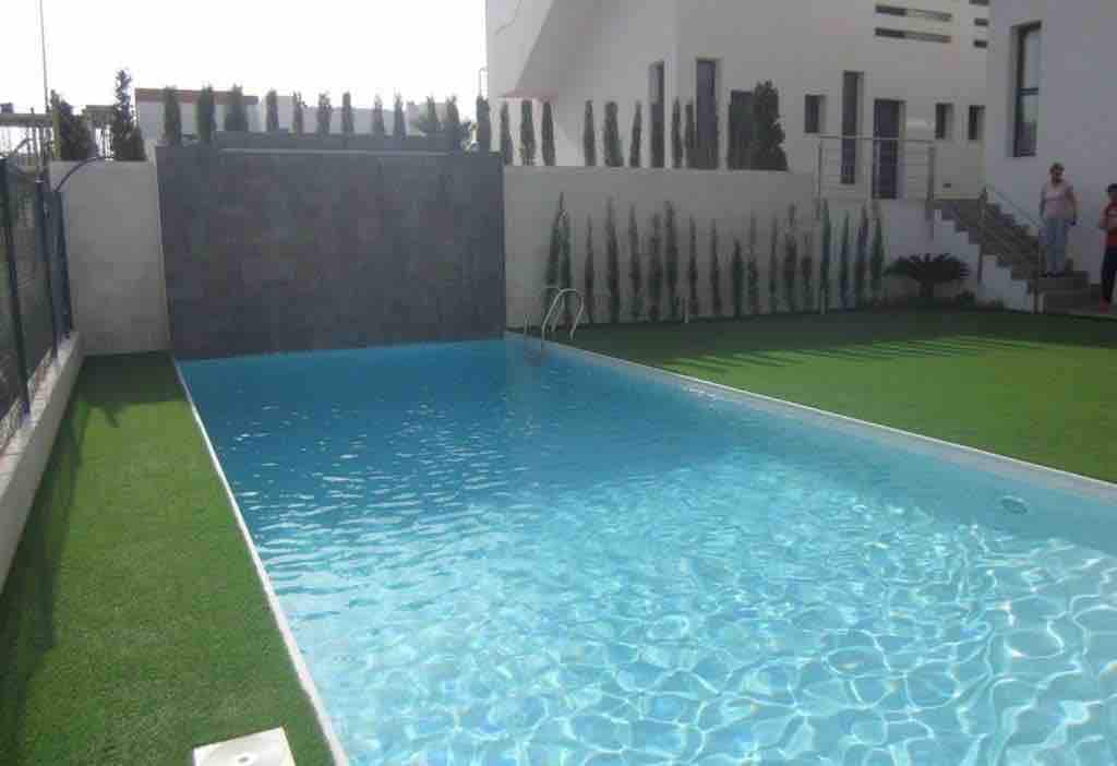 A large shared swimming pool is visible, featuring clear blue water that reflects sunlight. The pool area is surrounded by neatly trimmed grass and lined with tall, slender shrubs. A modern building can be seen in the background, enhancing the serene environment.