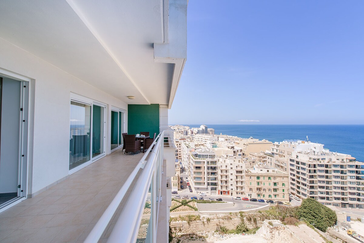 A spacious seafront terrace is visible, offering outdoor furniture and stunning views of the coastline. The balcony extends along the building, allowing for an unobstructed sight of both the sea and the nearby urban landscape. The clear blue sky complements the serene setting.