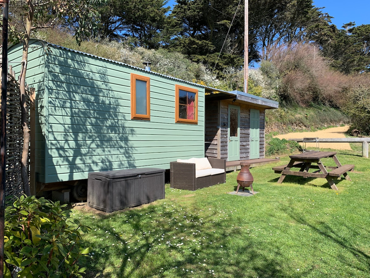 The exterior of a charming shepherd’s hut is displayed, featuring a green facade with wooden accents. A private garden area is highlighted, showcasing a picnic table and comfortable seating. The surrounding landscape includes grassy patches and natural foliage, with trees in the background under a clear blue sky.