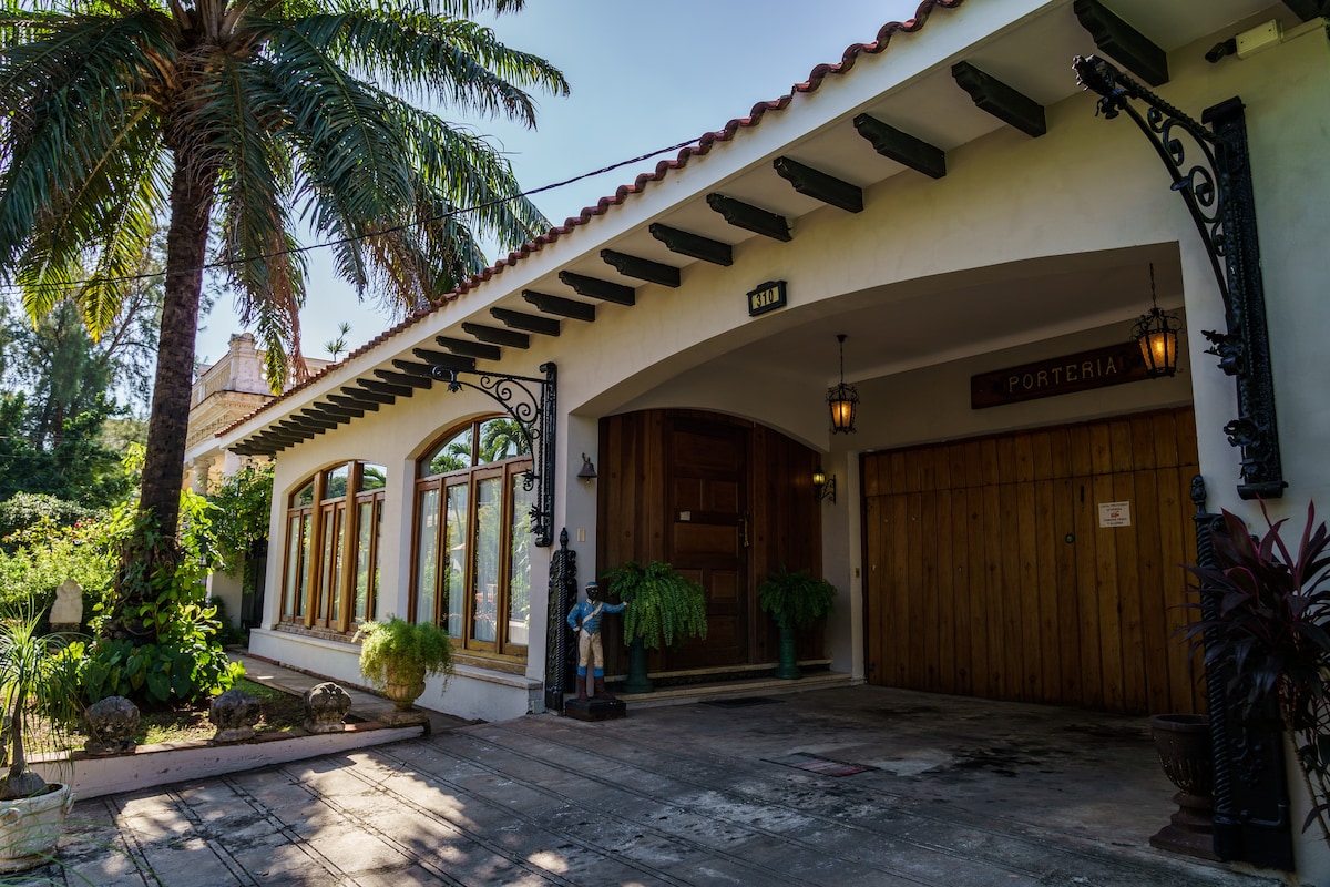 The entrance features an elegant façade with a wooden door and multiple arched windows, allowing natural light to fill the space. Lush palm trees and greenery are present in the surroundings, contributing to a serene atmosphere, complemented by decorative exterior lighting and artistic elements.