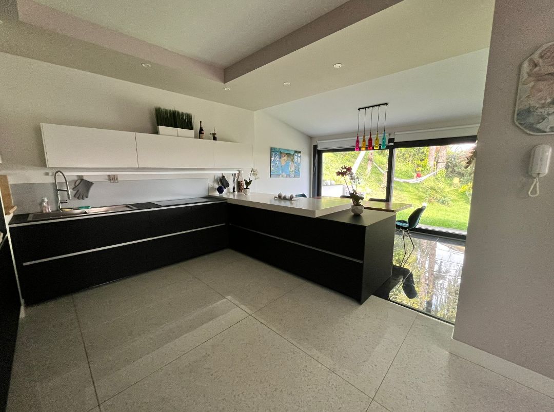 A modern kitchen is equipped with sleek cabinetry and a spacious island. Natural light flows in through large glass doors, leading to a green outdoor area. Colorful pendant lights hang above the island, complementing the minimalist design and creating a functional cooking space.