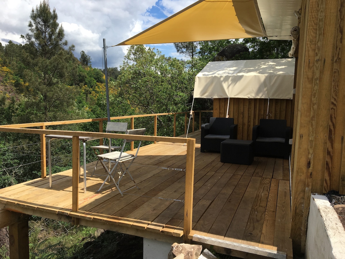 An outdoor deck is presented with wooden flooring, featuring a shaded area with a canopy. Two black seating options are provided alongside a small folding table and chair, creating a space for relaxation amidst the surrounding greenery.
