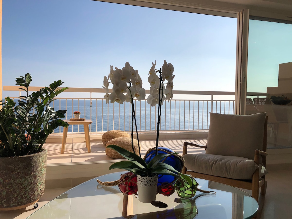 A bright living area features a glass table adorned with a potted orchid. A comfortable chair is positioned nearby, overlooking a panoramic view of the sea through large sliding glass doors, complemented by a serene coastal setting.
