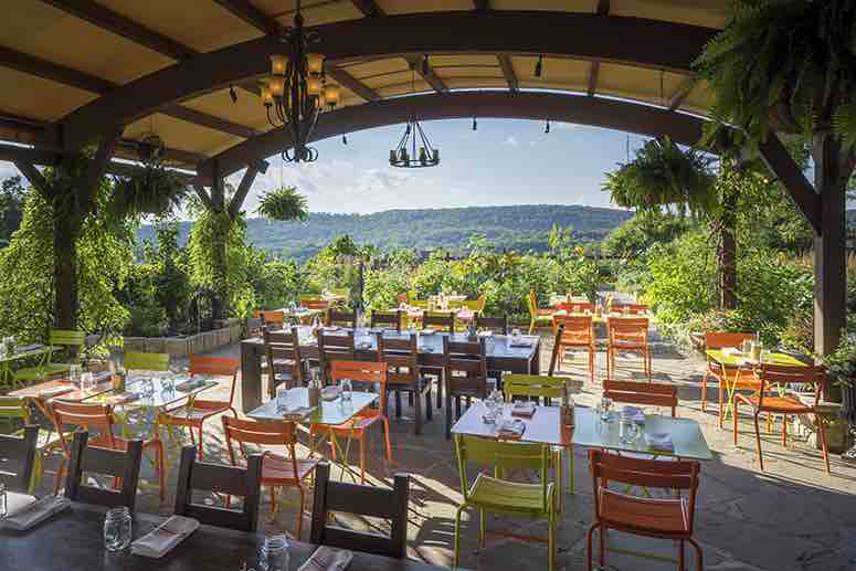 An outdoor dining area is framed by a wooden pergola, featuring colorful chairs in shades of orange and green. Tables are set with glassware, and lush greenery surrounds the space, offering panoramic views of the hills in the background under a clear blue sky.