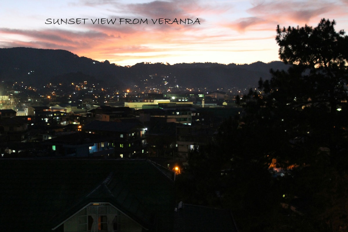 A sunset view is presented from the veranda, showcasing a vibrant sky filled with hues of orange and purple. Below, the town's lights begin to twinkle as dusk settles, forming a serene backdrop against the silhouette of distant mountains.