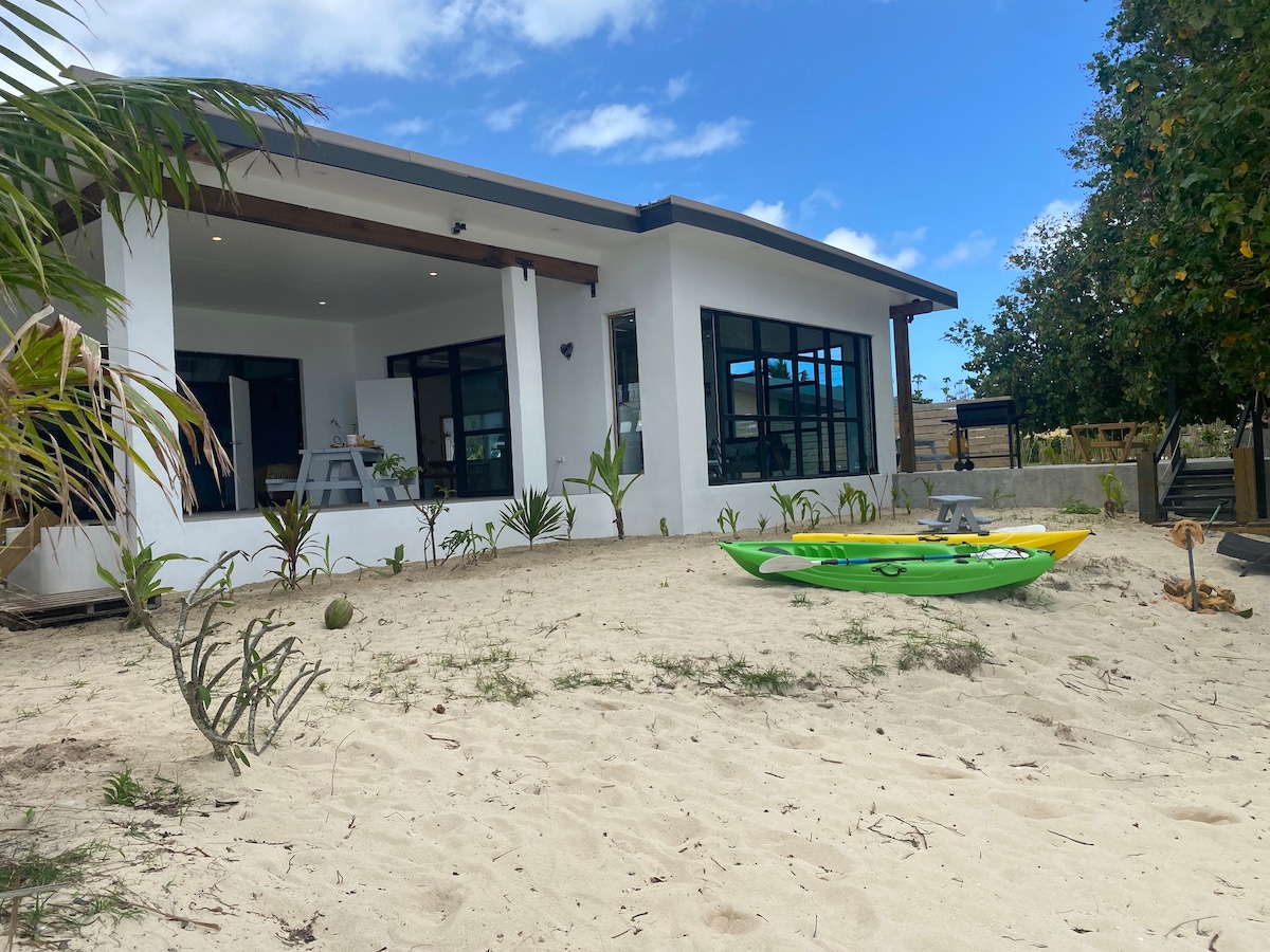 The beachfront house features a modern exterior with large glass doors opening to the living area. Kayaks in vibrant colors rest on the sandy ground, surrounded by lush green plants. Clear blue skies and soft clouds complete the serene tropical setting.