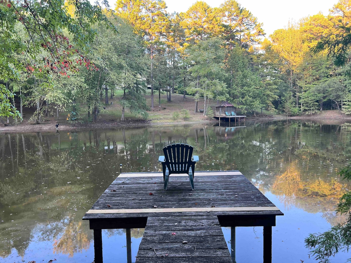 A wooden dock extends over a calm pond, featuring a single green Adirondack chair. Surrounding trees reflect softly in the water, providing a serene backdrop. A small boathouse is visible on the far side, adding to the peaceful atmosphere of the property.