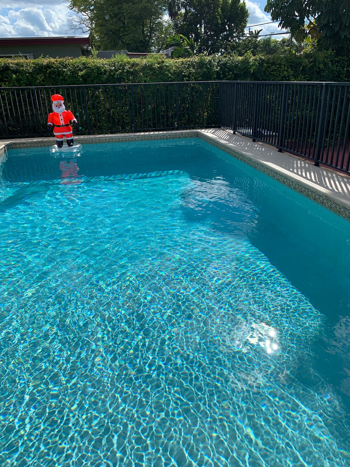 A private pool is surrounded by a black metal fence, with a clear, bright blue water surface reflecting sunlight. A decorative Santa figure stands on the pool's edge, adding a festive touch to the tranquil outdoor space.