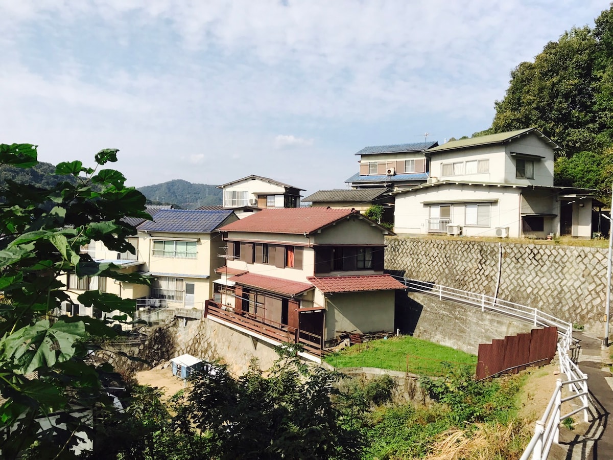 A collection of residential buildings is visible on a hillside, showcasing a blend of architectural styles. The structures are surrounded by greenery and accessed via sloped terrain, with some featuring red roofs. A clear sky provides a bright backdrop for this tranquil setting.