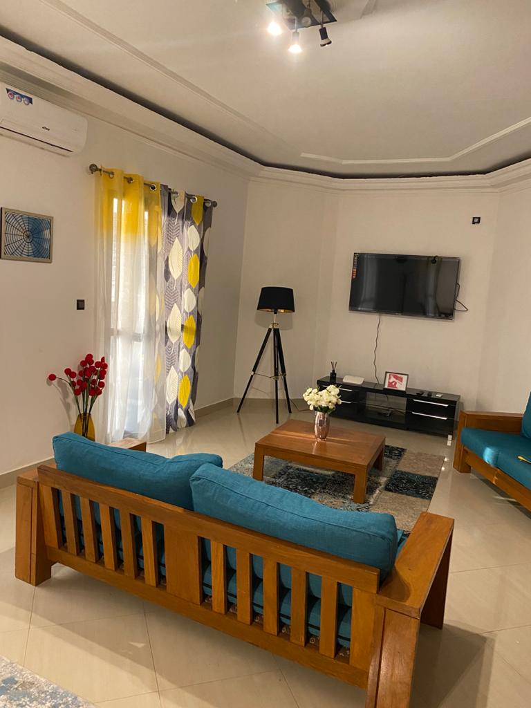 A bright living area features a wooden seating arrangement with turquoise cushions. A coffee table sits centrally, surrounded by decorative elements. A modern television is mounted on the wall, with natural light filtering through curtains adorned with geometric patterns.
