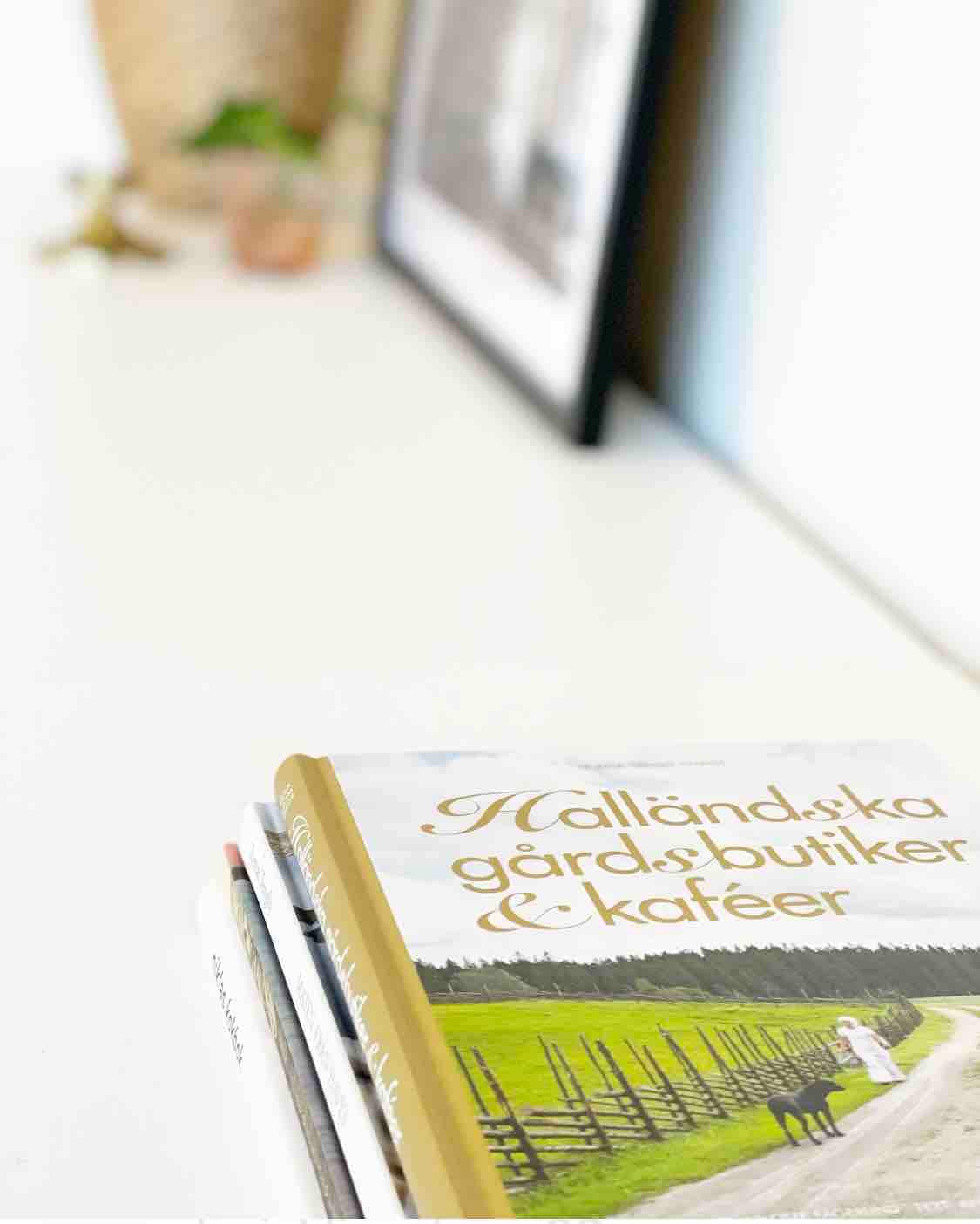 A clean, minimalistic tabletop is shown with a small stack of books, the top one featuring the title 'Halländska gårdsbutiker & kaféer.' In the background, a plant and a framed picture add subtle decor elements to the space.