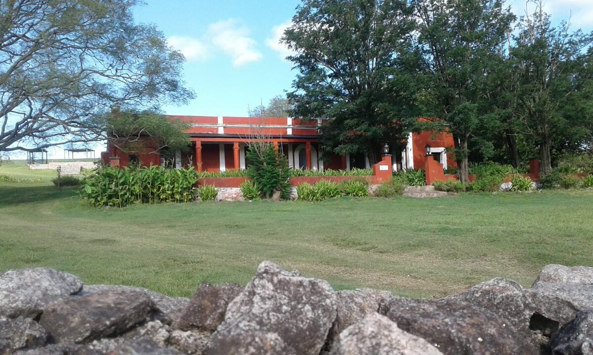A rustic casona is seen, surrounded by lush greenery and trees. The structure features red-toned walls, with a porch that opens to a landscaped area. A stone wall is present in the foreground, adding a natural element to the tranquil setting.