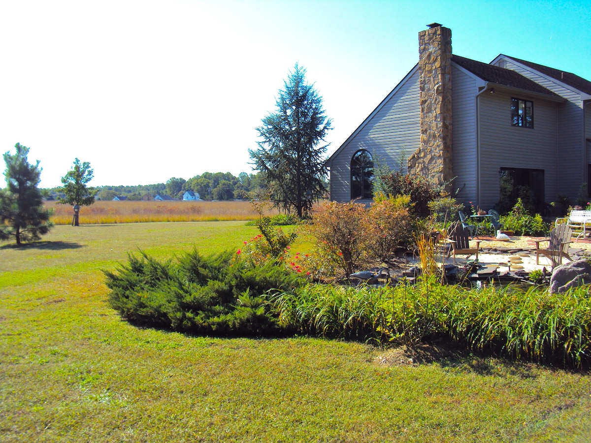 A spacious yard is bordered by lush greenery and colorful shrubs, leading to a modern home featuring a prominent stone chimney. Open fields extend in the background, offering a peaceful view of the natural landscape.