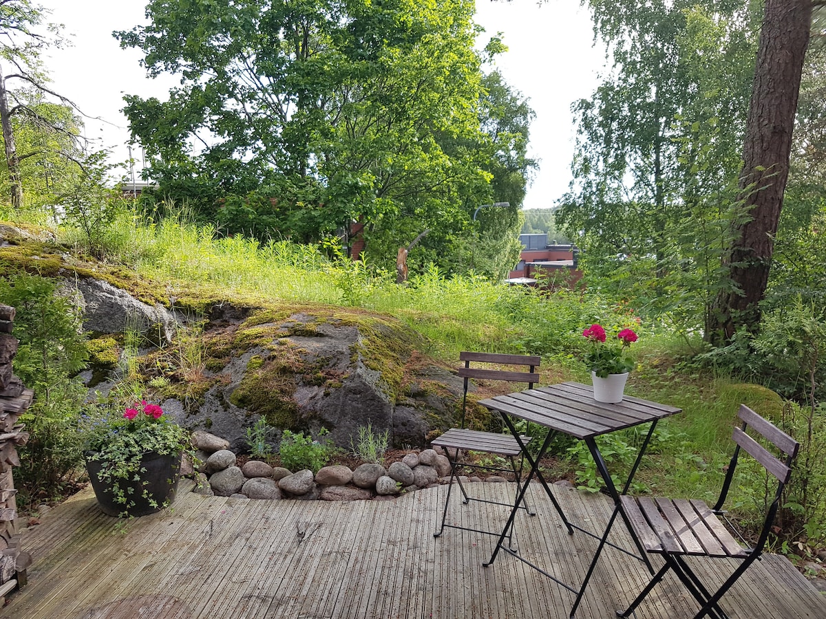 A small backyard area is depicted, featuring a simple metal table with two folding chairs. A potted plant with vibrant flowers sits on the table. Lush grass and rock formations create a natural backdrop, surrounded by trees that enhance the serene outdoor environment.