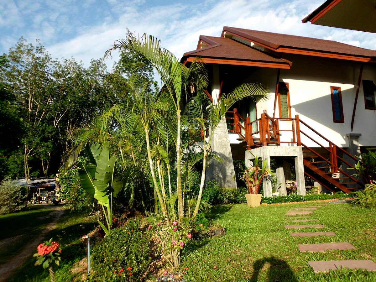 A well-maintained exterior of a villa is surrounded by lush greenery and vibrant flowers. Steps lead to a welcoming entrance, while tropical plants add natural beauty to the landscape. The structure features a pitched roof and large windows, allowing ample light into the home.
