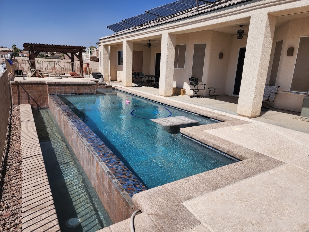 An infinity pool glistens under the sun, flanked by a spacious patio area featuring seating arrangements. Nearby, a gazebo provides shade, and solar panels are visible on the roof. The clear water reflects the blue sky, creating a serene outdoor environment.