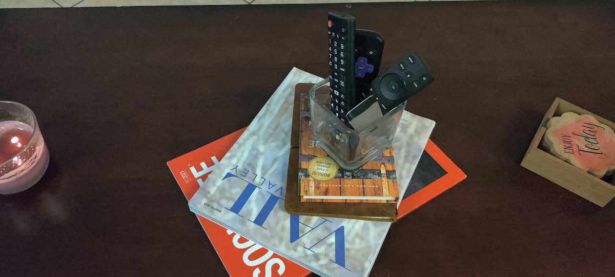 An organized coffee table displays a variety of items, including magazines stacked in an artistic arrangement. A decorative holder contains four remote controls, while a candle casts a warm glow in the background. A small wooden box with the word 'Teddy' is also present.