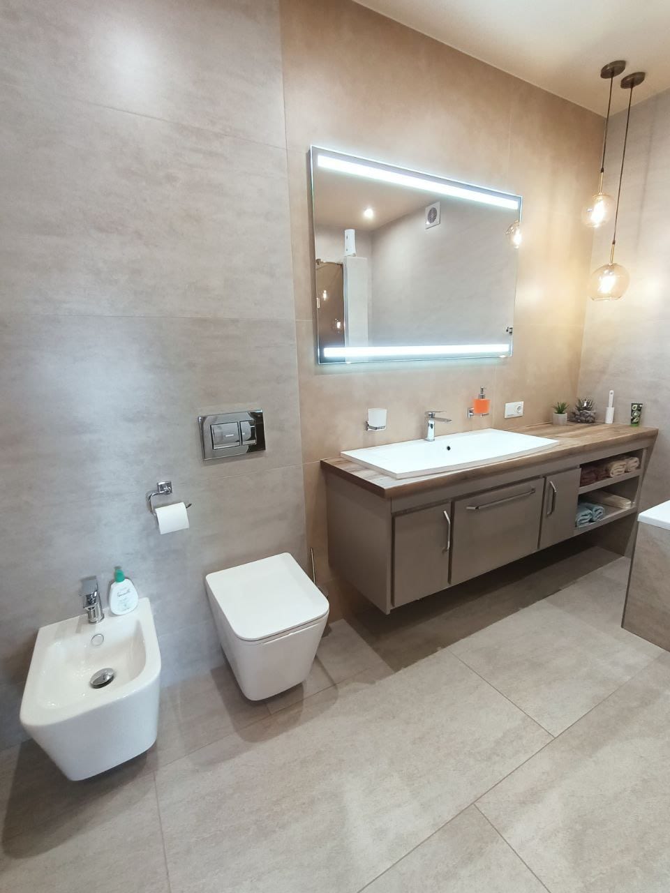 A modern bathroom is featured, showcasing a sleek vanity with a lighted mirror above. The space includes a white toilet and bidet, with neutral-toned tiles and warm lighting that enhances the serene environment.