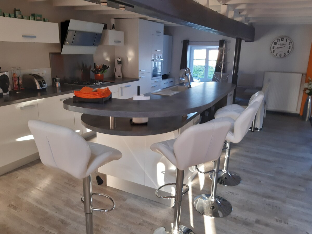 The kitchen area features a modern, streamlined design with a large central island and four high stools. Light-coloured cabinetry is complemented by stainless steel appliances. A wall clock and bright windows provide a sense of openness and natural light in the space.