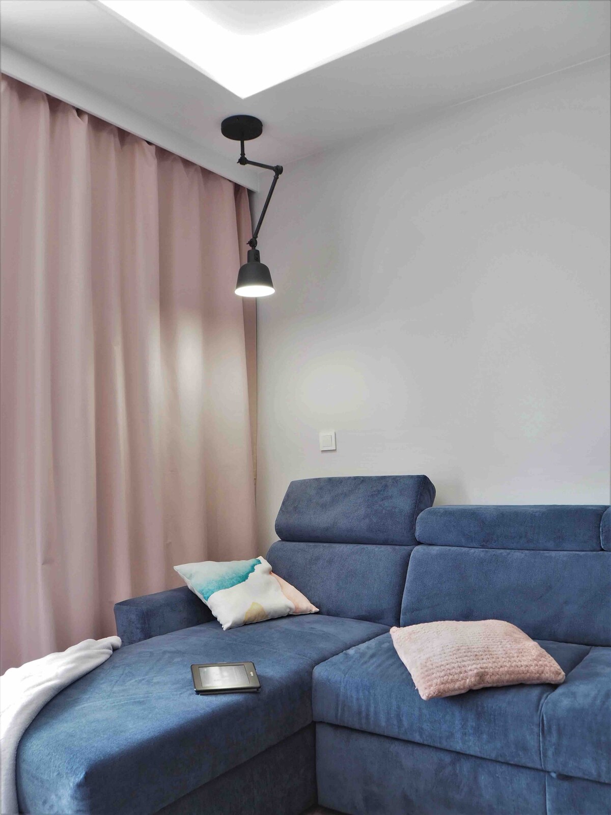 A corner sofa in a soft blue tone is complemented by light pink curtains that add warmth to the space. A simple pendant light illuminates the area, while cushions in various colors are arranged on the sofa, enhancing comfort without overwhelming the design. A cozy blanket rests nearby.