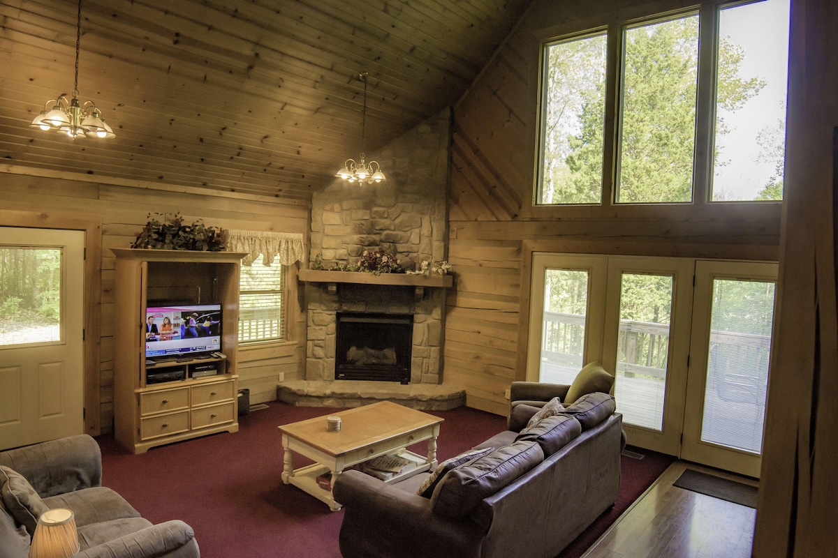 A spacious living area features a high, wooden ceiling and large windows offering views of the surrounding nature. A stone fireplace is centrally located, with a TV mounted above it. Comfortable couches are arranged around a light wooden coffee table, creating a welcoming space.
