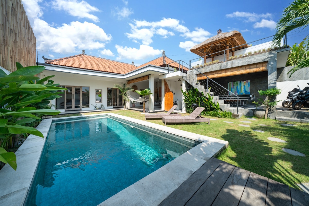 A spacious villa features a large swimming pool surrounded by lush greenery. Comfortable sunbeds are positioned on the pool deck. The covered terrace showcases wooden accents and easy access to the interior spaces, all under a bright blue sky with scattered clouds.