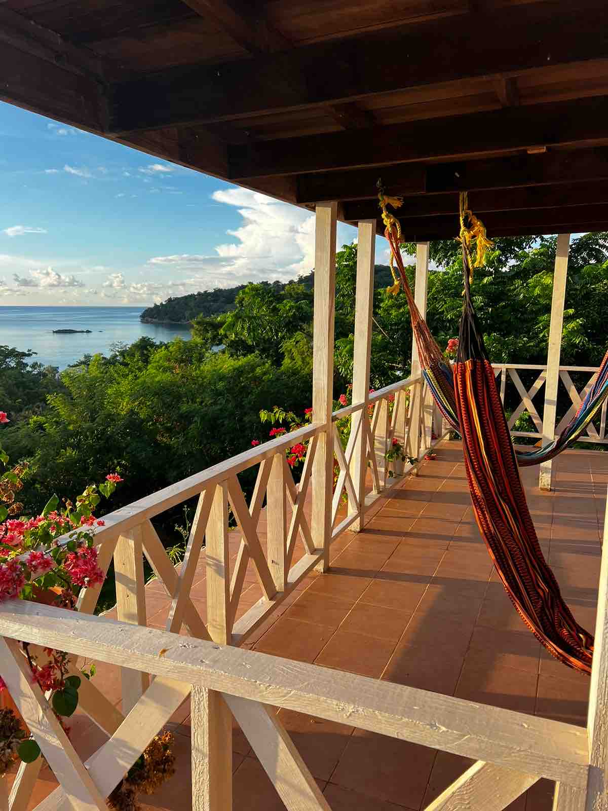 A terrace is showcased with wooden beams overhead and colorful hammocks suspended between posts. Lush greenery and bright flowers frame the view, which extends to the serene ocean and distant coastline under a clear sky.