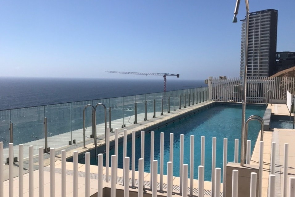 An outdoor swimming pool area is visible, featuring a clear blue water surface that reflects the sky. The pool is framed by a glass barrier and white railings, offering views of the ocean and surrounding buildings. A construction crane can be seen in the distance.