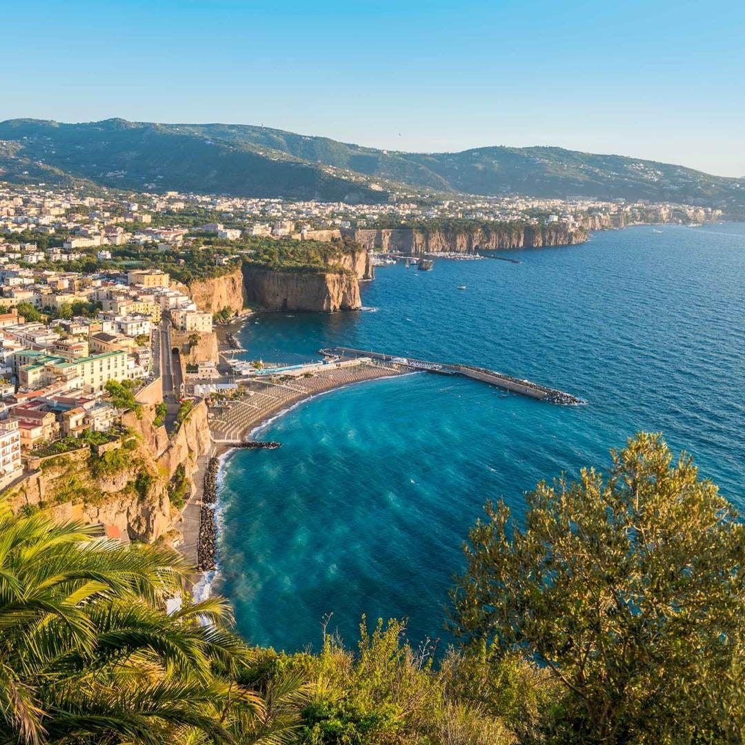 The image captures a coastal landscape featuring a serene bay embraced by cliffs and a town nestled along the coastline. Clear turquoise waters reflect the sky, while lush greenery frames the scene, highlighting the beautiful natural surroundings.