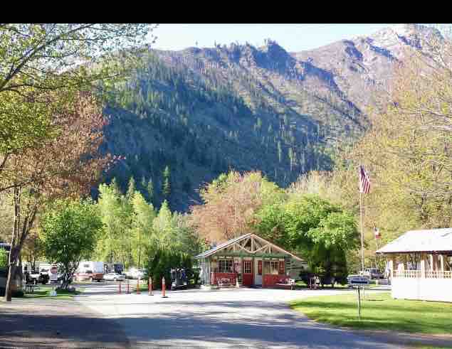 A welcoming entrance to a campground is framed by lush greenery and towering mountains in the background. A rustic office building is visible, along with a well-maintained road leading into the park. Seasonal trees display hints of color, enhancing the natural setting.
