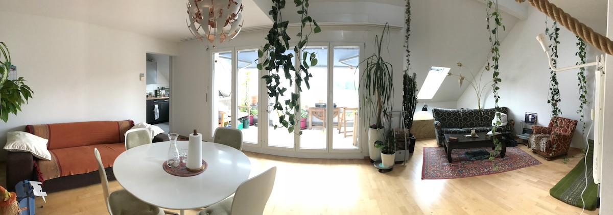 A spacious living area features large glass doors that open to a balcony, allowing natural light to fill the room. Comfortable seating options are arranged alongside decorative plants. A round dining table sits nearby, inviting shared meals and gatherings.